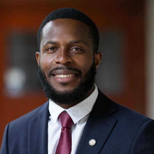 A smiling Black man with a beard and short hair wears a navy suit, polka dot shirt, and burgundy tie, with a Notre Dame lapel pin.